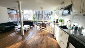 A modern kitchen with white cabinets, a black countertop, and a stainless steel sink. There is a dining area with a round table and chairs, and a black sofa.
