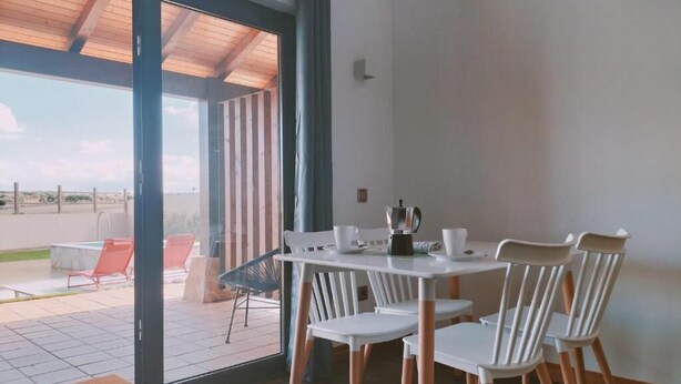 Een eetruimte met een tafel en stoelen, een koffieapparaat en uitzicht door een glazen deur op een terras met stoelen.