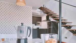Een moderne keuken met een koffiezetapparaat, een schaal fruit en een glas yoghurt op een tafel. In de achtergrond is een trap met houten treden en een metalen leuning zichtbaar.