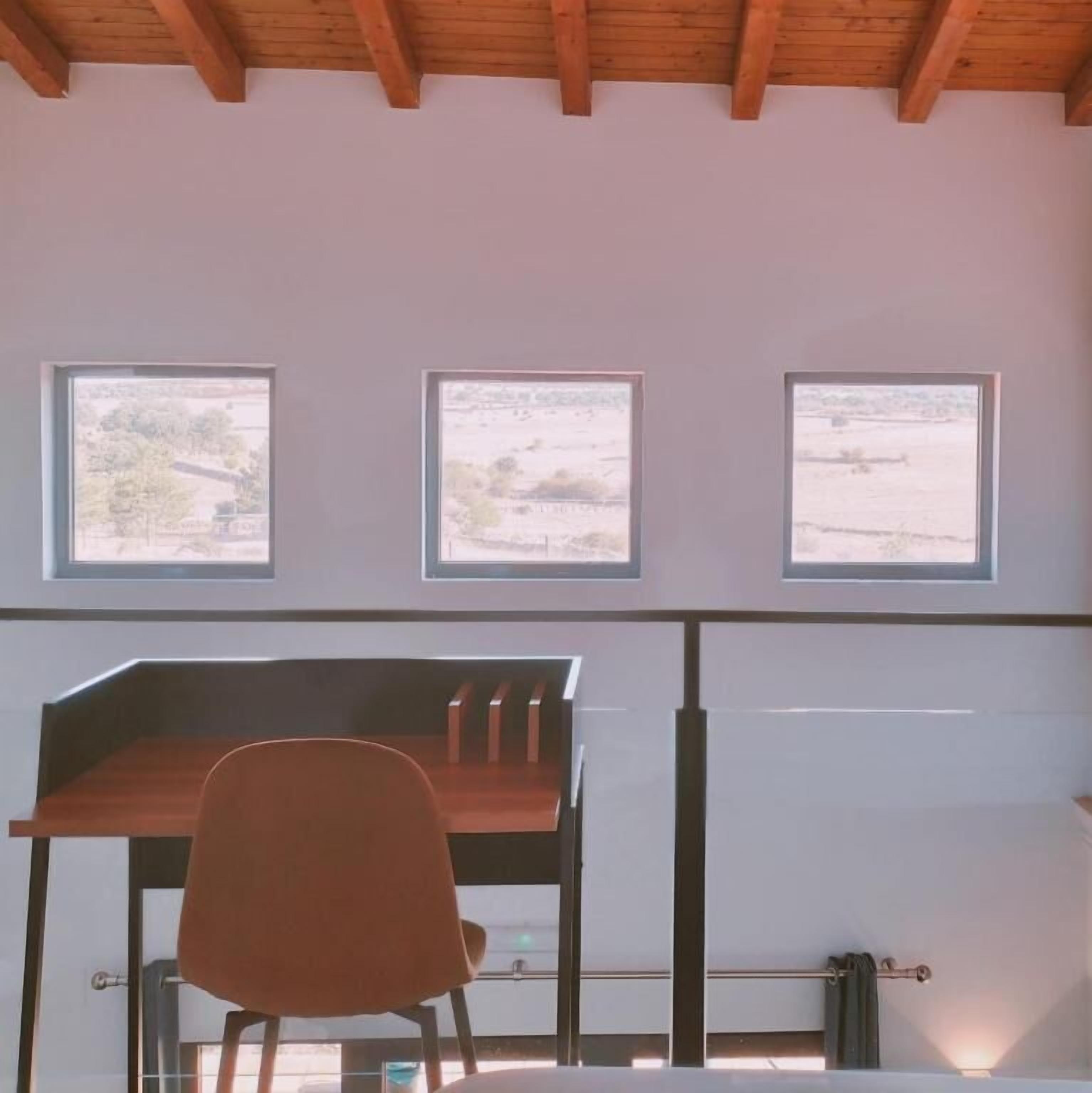 A room with a wooden desk, a chair, and three windows providing views of the outdoors.