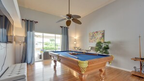 Ein Billardtisch in einem Raum mit Holzboden, Deckenventilator, einer Couch und einem großen Fenster mit Vorhängen.