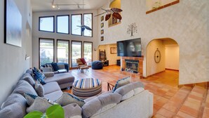 Ein geräumiges Wohnzimmer mit einer großen Eckcouch, einem runden Hocker und einem Kamin. Mehrere Fenster sorgen für natürliches Licht, und ein Deckenventilator ist installiert.