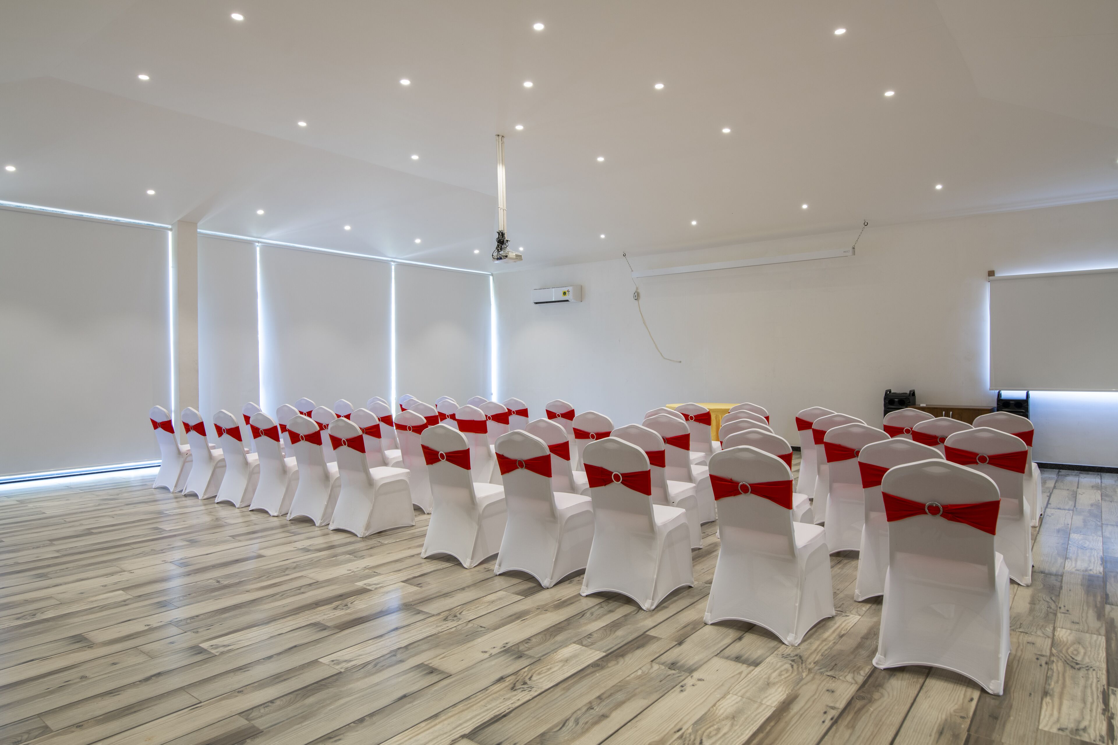 A conference room with rows of white chairs with red sashes, wooden floors, and recessed ceiling lights.