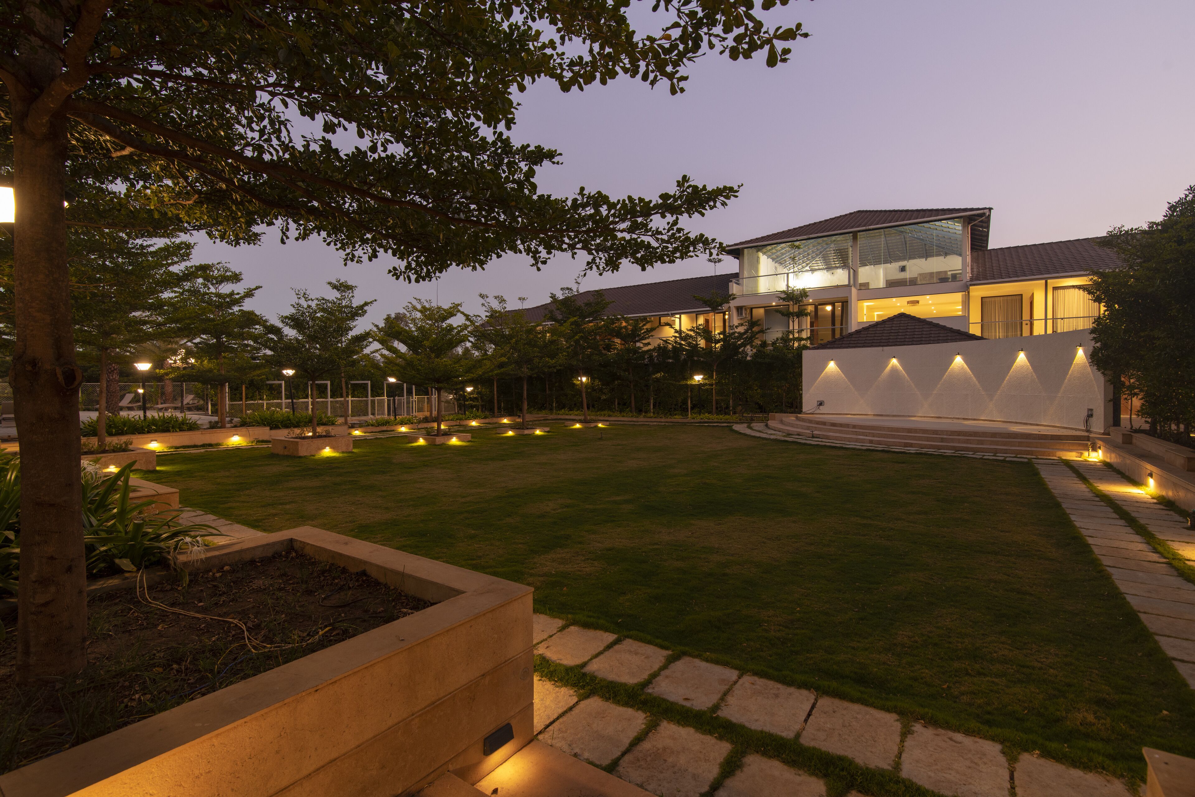 Un bâtiment moderne à la façade vitrée, entouré d’un jardin bien éclairé, agrémenté de sentiers et de jeux de lumière.