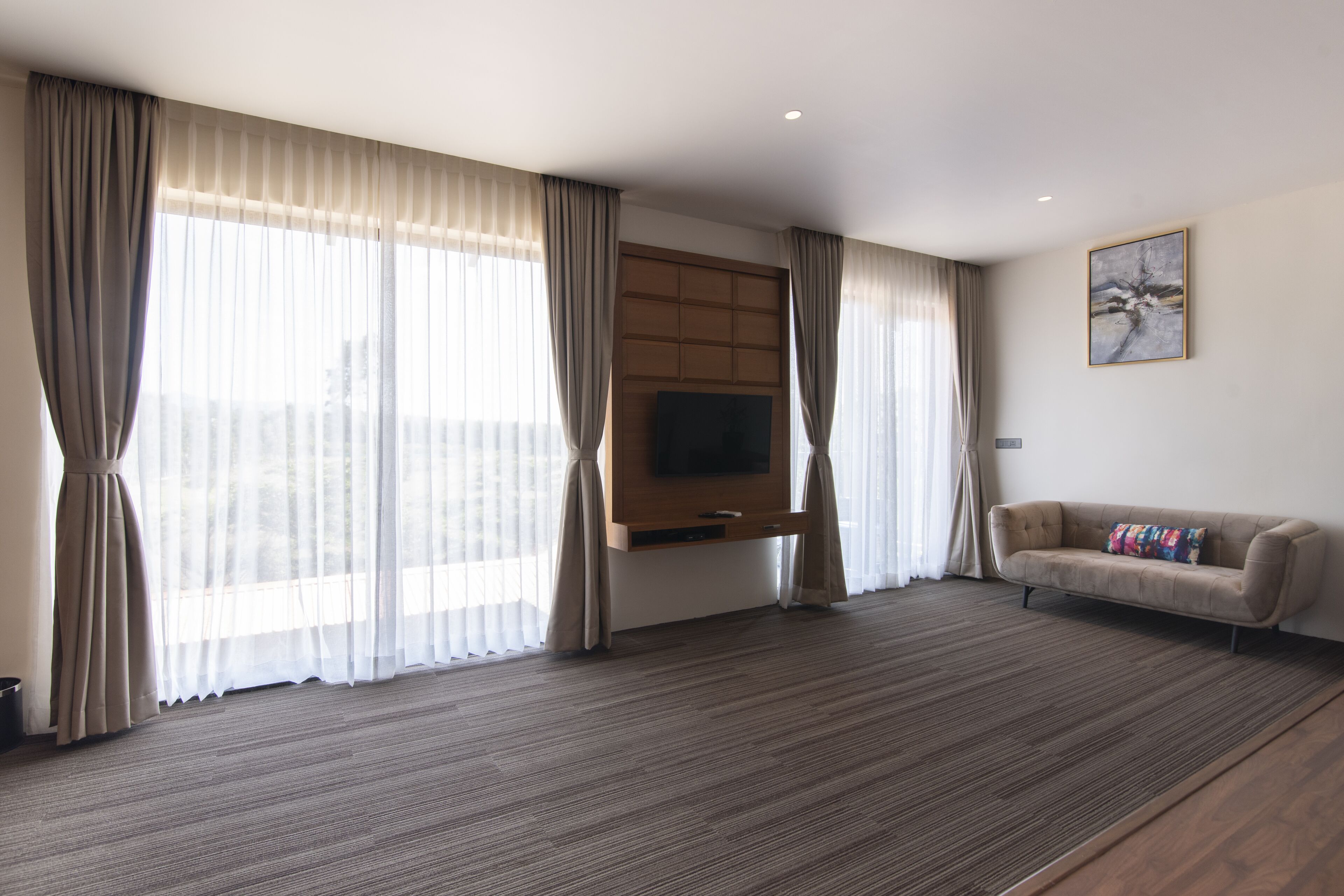 A modern hotel room with a large window, a flat-screen TV mounted on the wall, a sofa, and a carpeted floor.