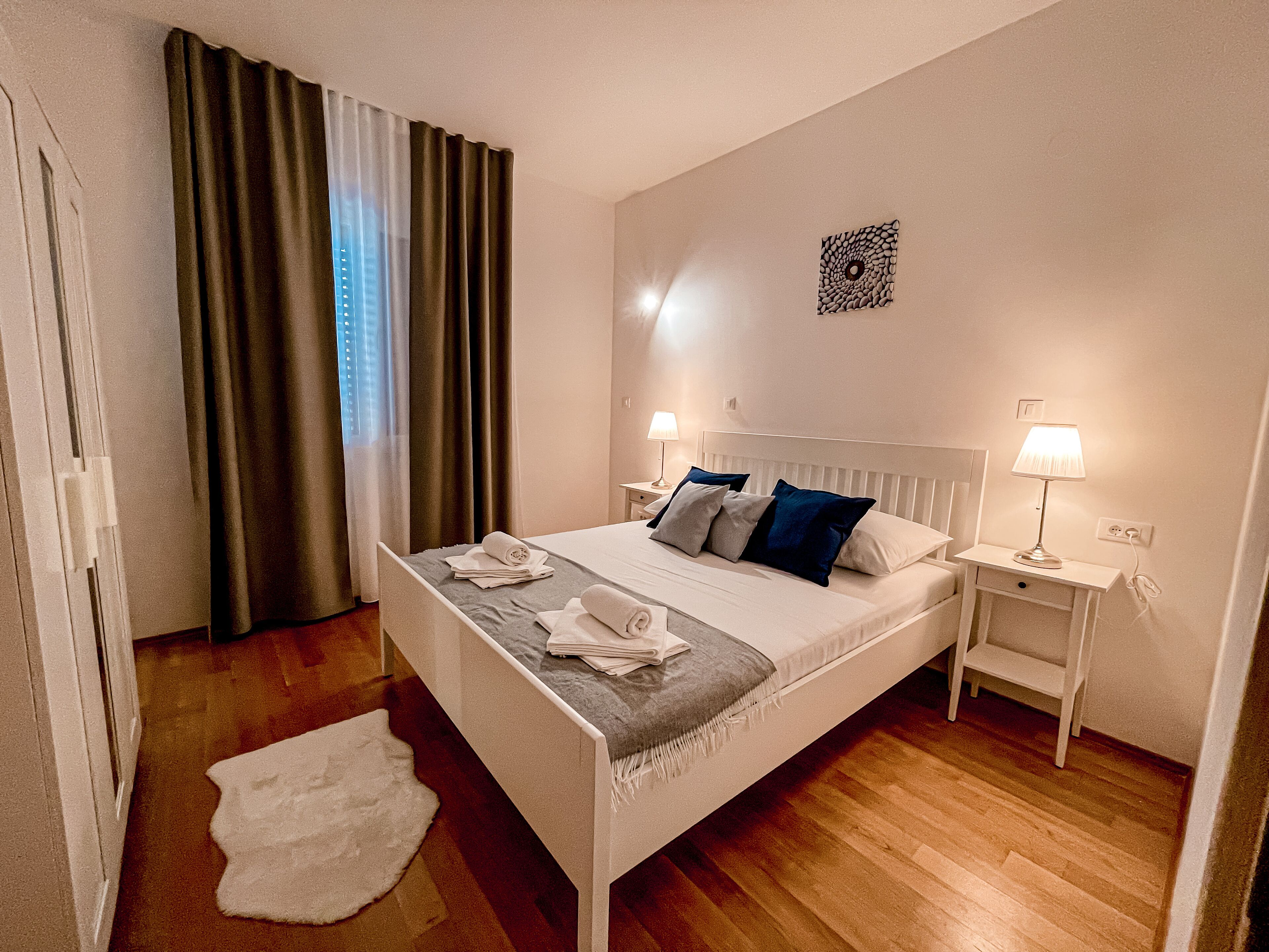 Una habitación de hotel ordenada con una cama, dos mesitas de noche, una obra de arte en la pared y un ventanal con cortinas.