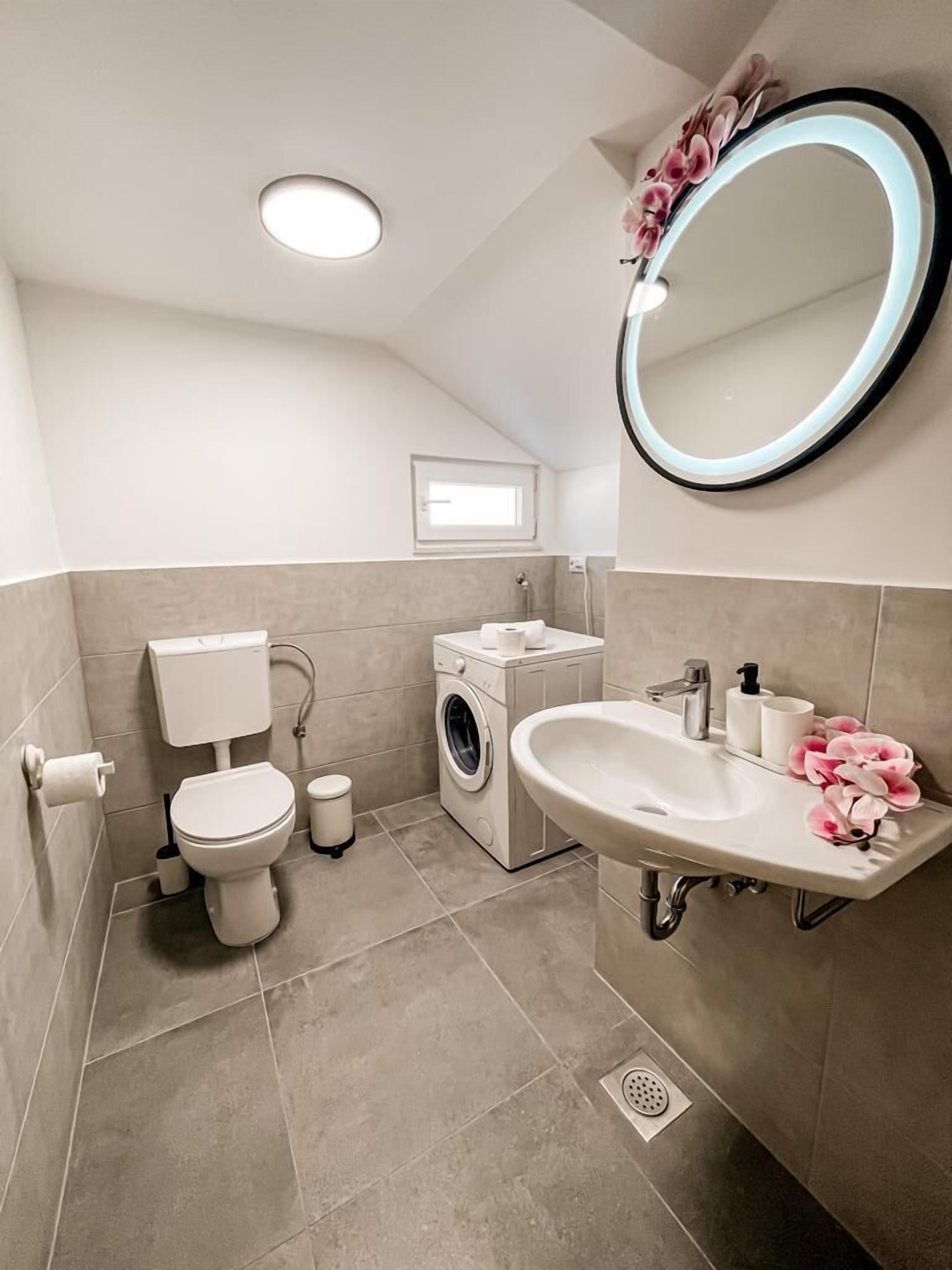 A modern bathroom with a toilet, washing machine, sink, and a mirror with a light.