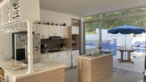 Una cocina moderna con una isla central, horno empotrado y refrigerador decorado con imanes. Se observa una ventana grande con vistas a una piscina y zona de estar exterior.