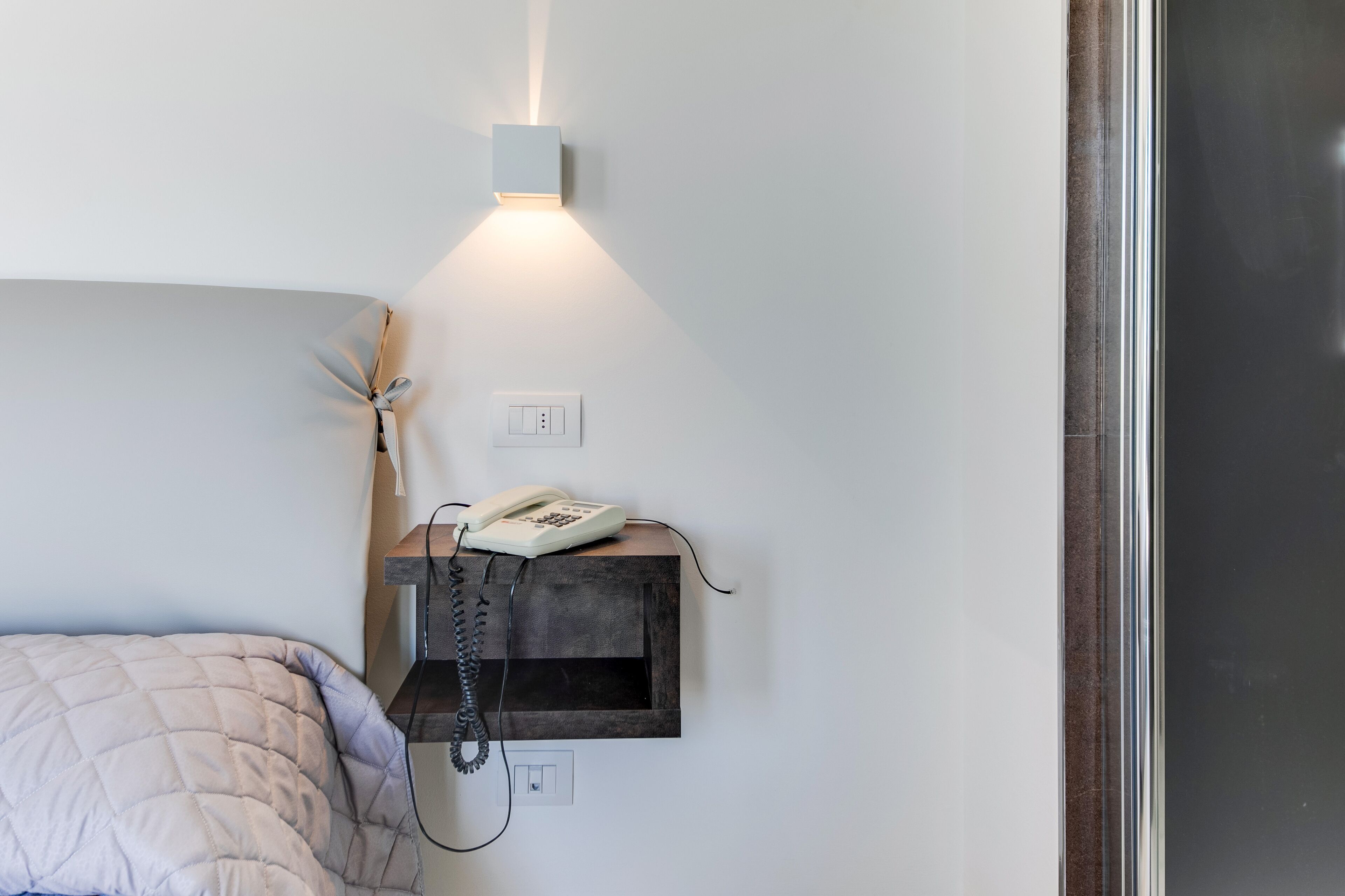 Een slaapkamer met een bed, een nachtkastje met telefoon en een wandlamp.