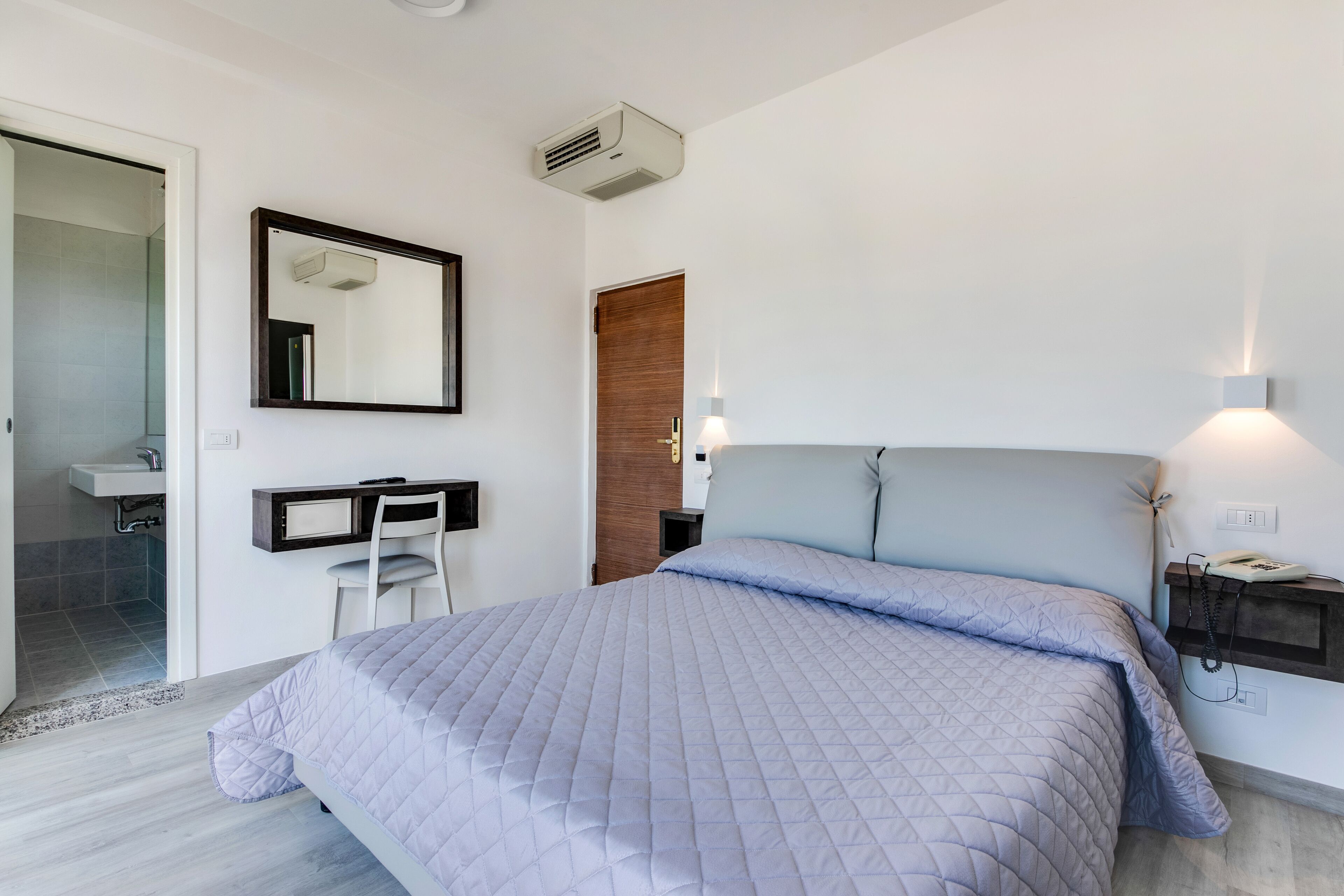 Quarto moderno com cama, escrivaninha, cadeira, espelho e mesa de cabeceira com um telefone.