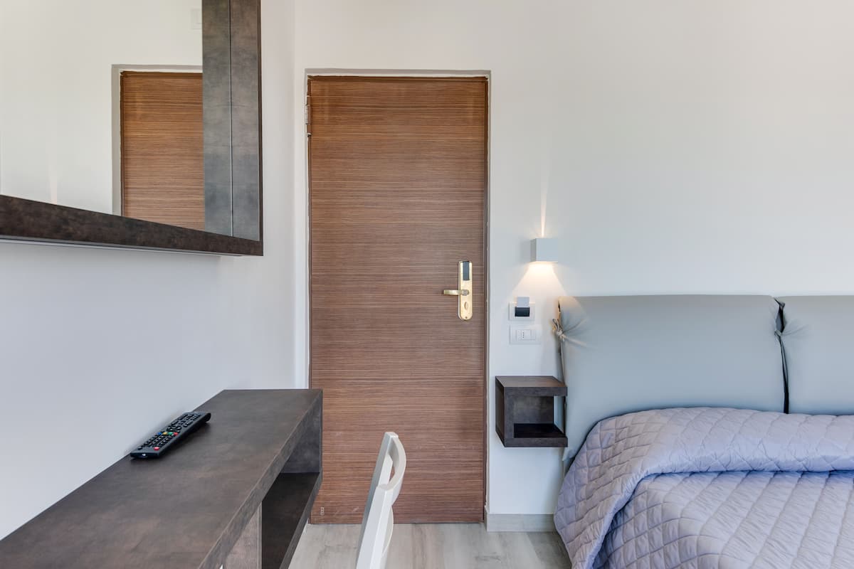 A hotel room with a wooden door, a bedside table with a remote control, a bed with a quilted cover, and a wall-mounted night light.