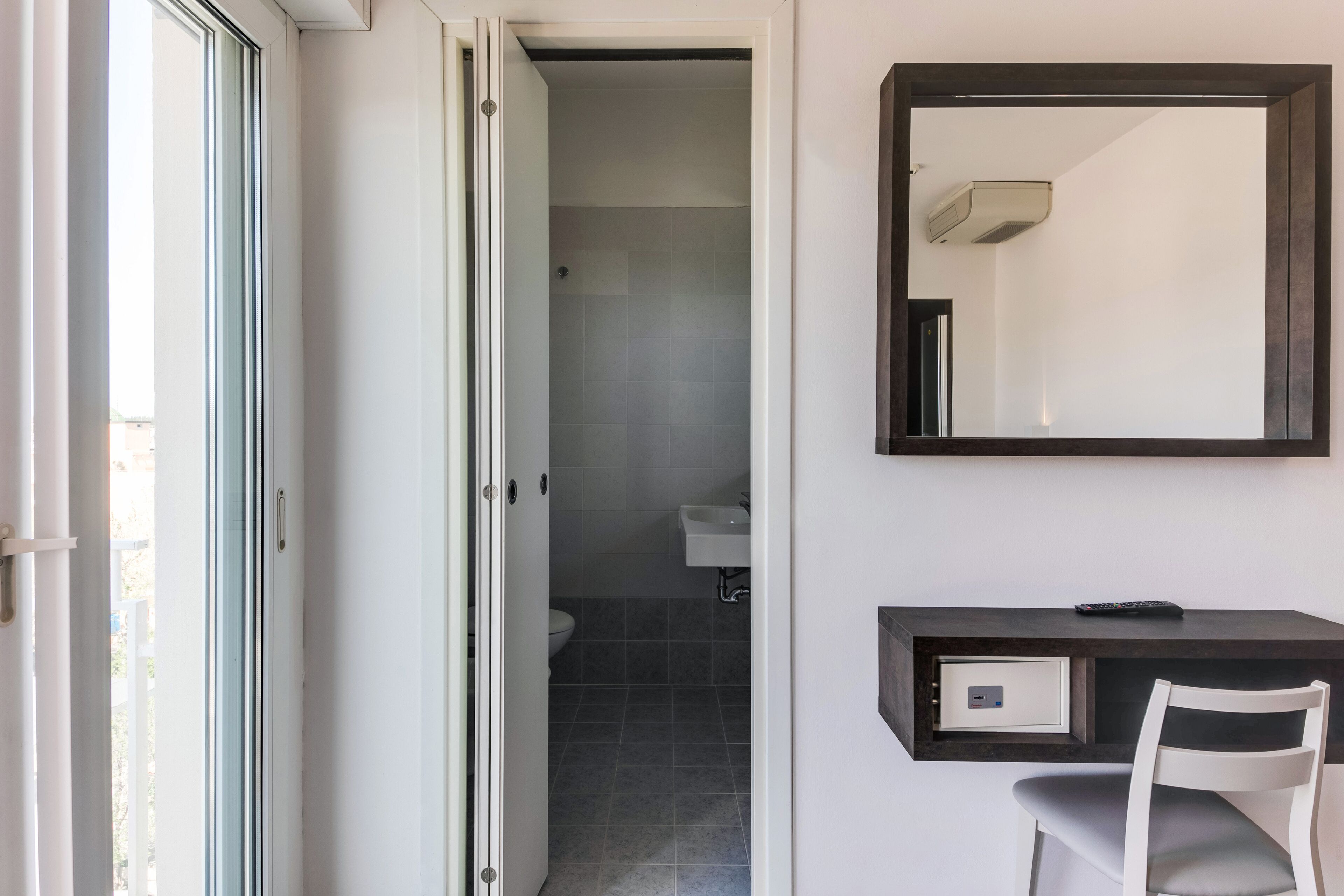 A bathroom with a toilet, sink, and shower. There is a white chair and a dark wooden desk with a remote control on it.