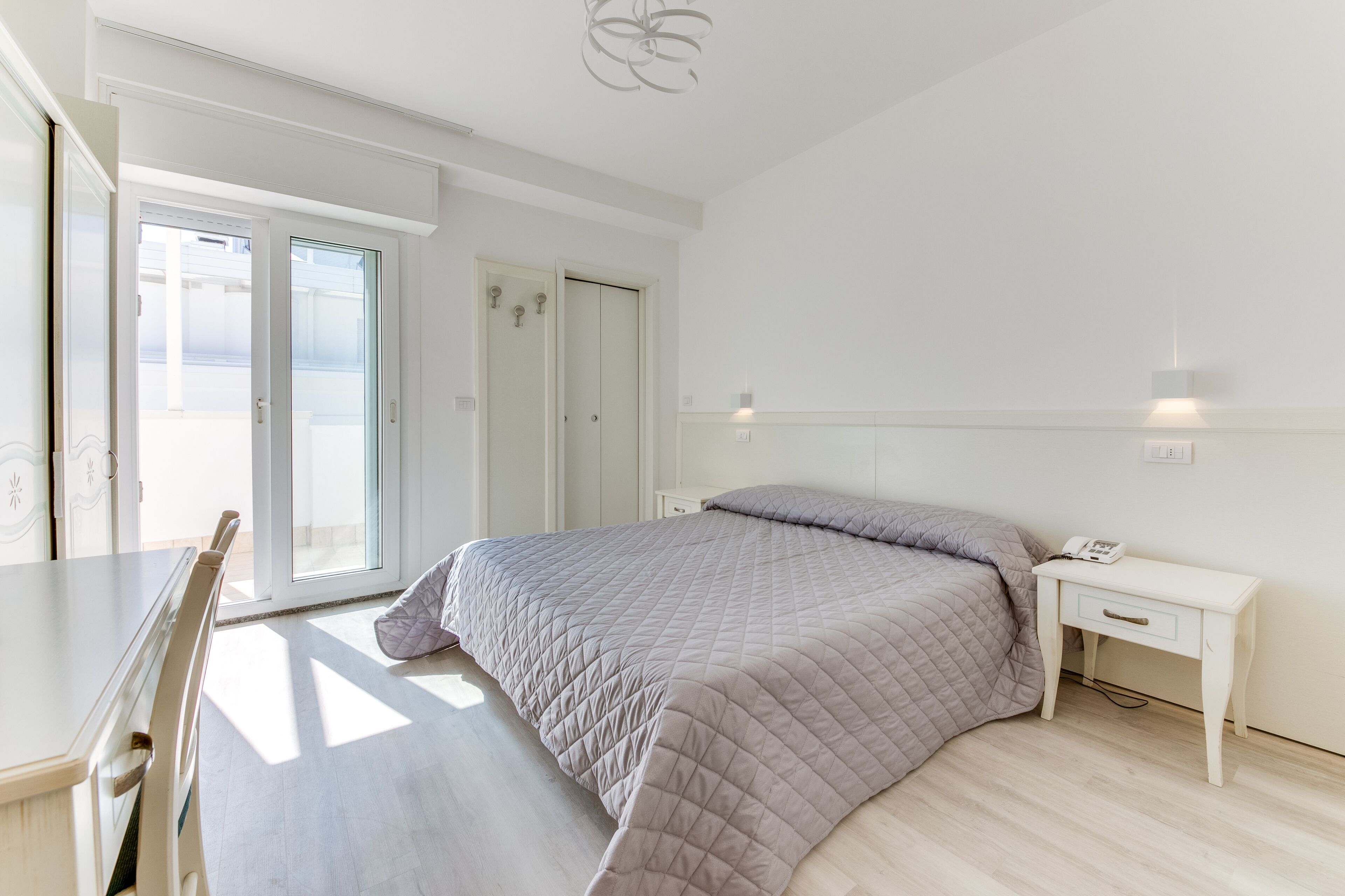 Quarto com cama, mesa de cabeceira, cadeira e porta de vidro deslizante para uma varanda.
