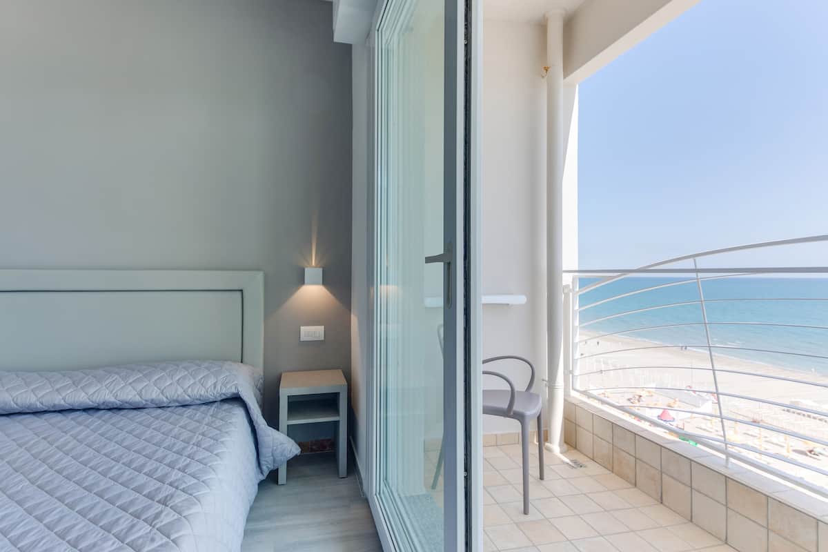 A bedroom with a bed, a nightstand, and a chair by a balcony with a view of the beach.