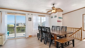 A dining area with a wooden table and chairs, a ceiling fan, and a view of the beach through large windows. - Skyview by the Sea (Corolla)