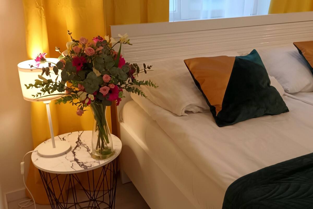 Una cama bien hecha con sábanas blancas, dos almohadas y una manta verde. Un florero con flores está sobre una mesita redonda al lado de la cama.
