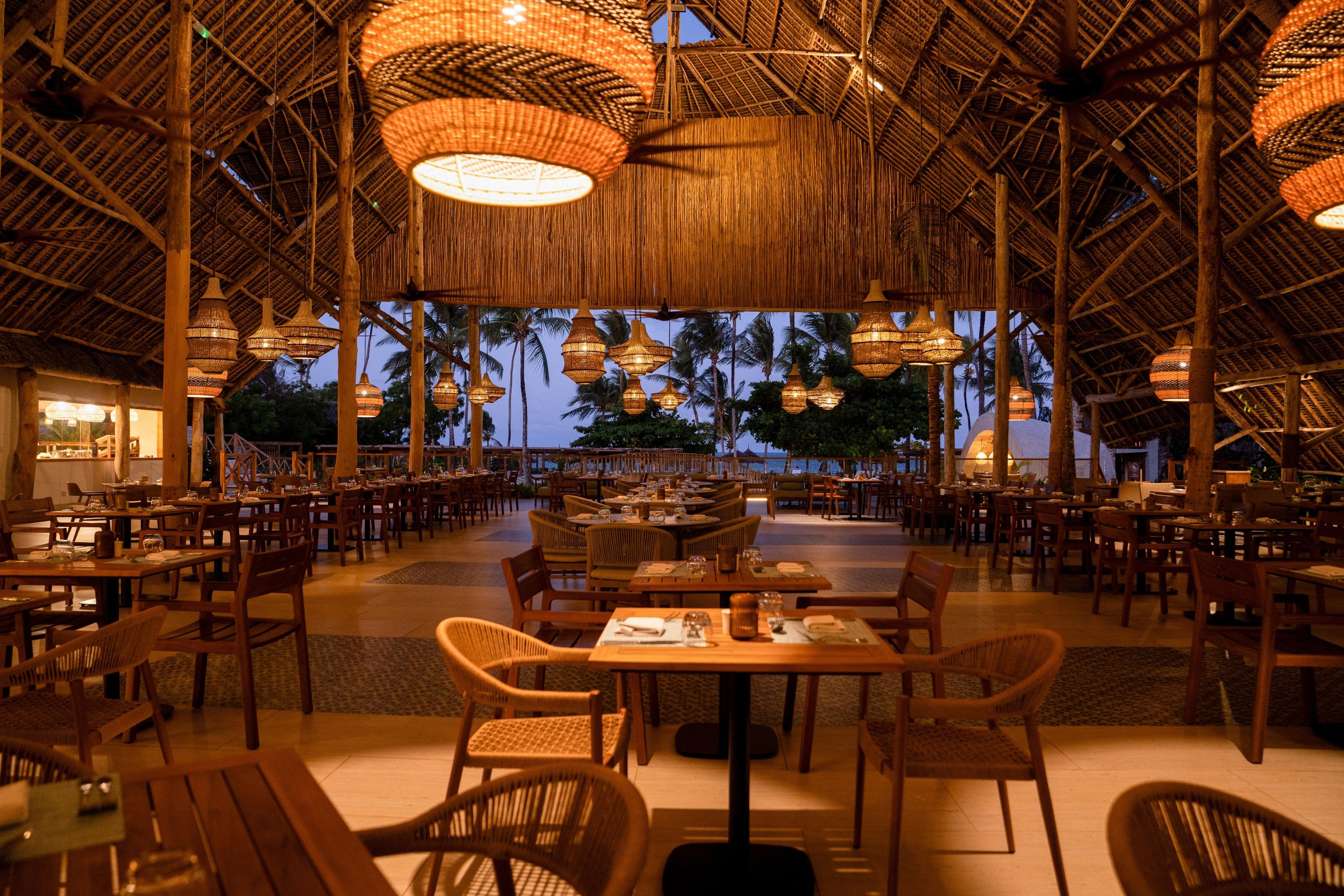 A spacious restaurant with high ceilings and thatched roof design, featuring multiple dining tables and chairs.