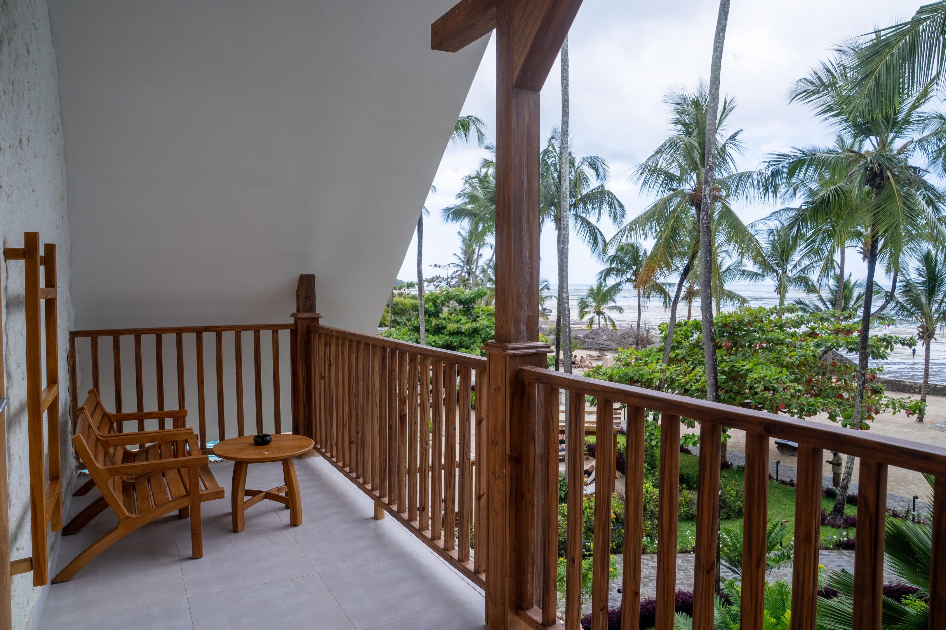 A balcony with wooden furniture, a view of the beach, and palm trees.