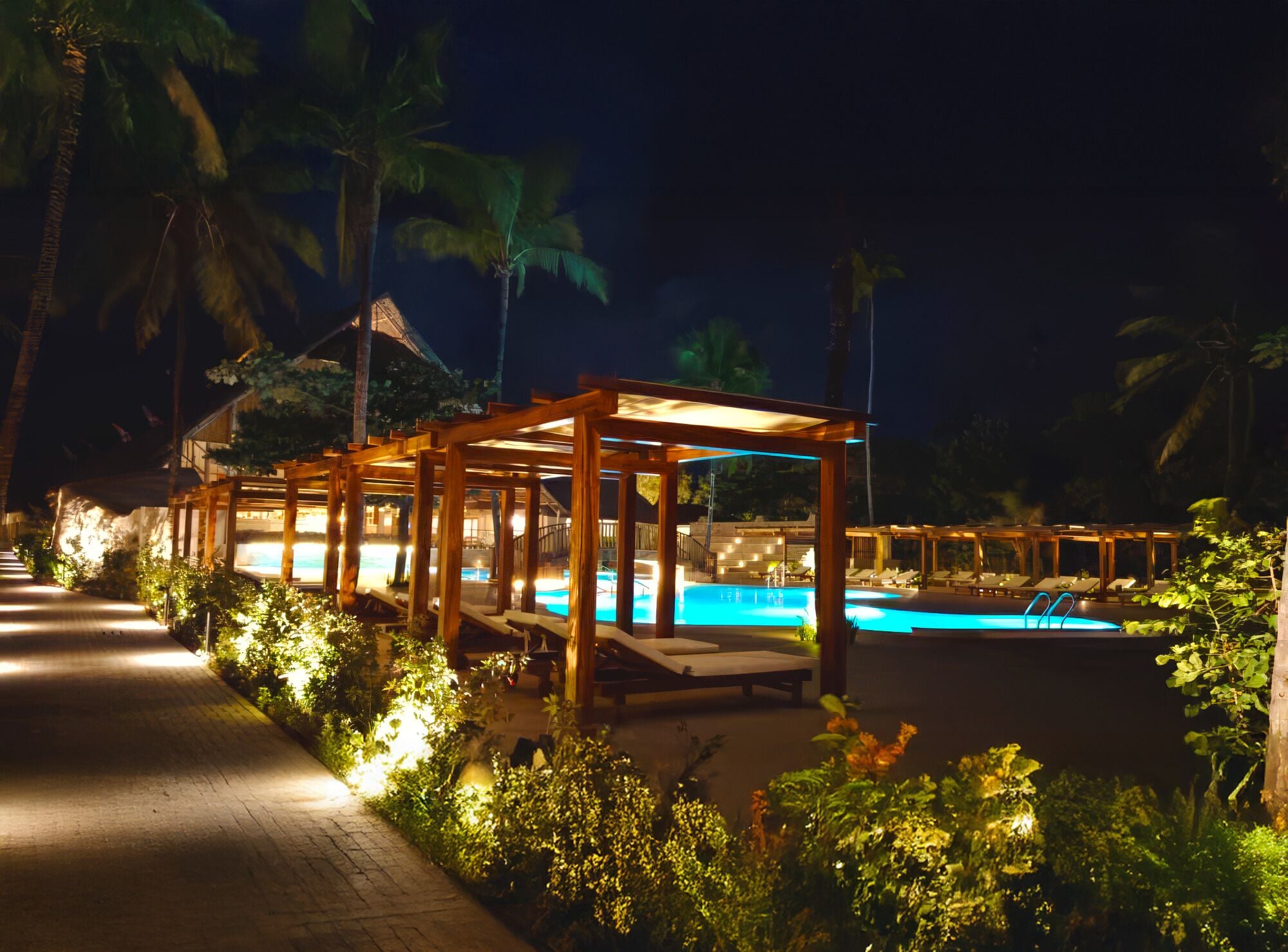 Zona de piscina en un resort nocturno con pérgolas de madera, sillas de descanso y un camino.