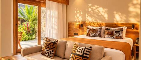 A hotel room with a bed, two sofas, a small table with a flower, and a view of greenery outside.