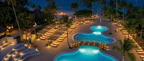 A pool area with lounge chairs and a curved pool, surrounded by palm trees and a beach view.