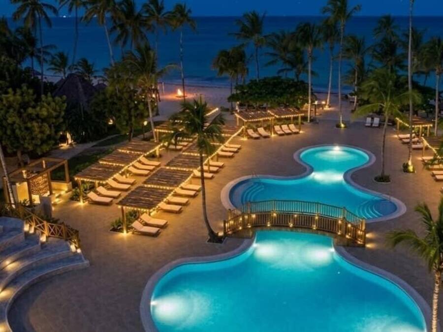 Ein Poolbereich mit Liegestühlen und einem geschwungenen Pool, umgeben von Palmen und Blick auf den Strand.