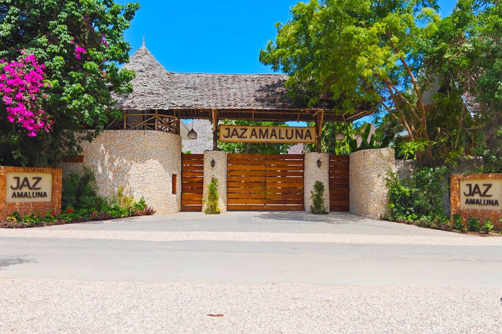 Una entrada porticada a una propiedad llamada Jaz Amaluna, con un muro de piedra, un portón de madera y una estructura con techo de paja.
