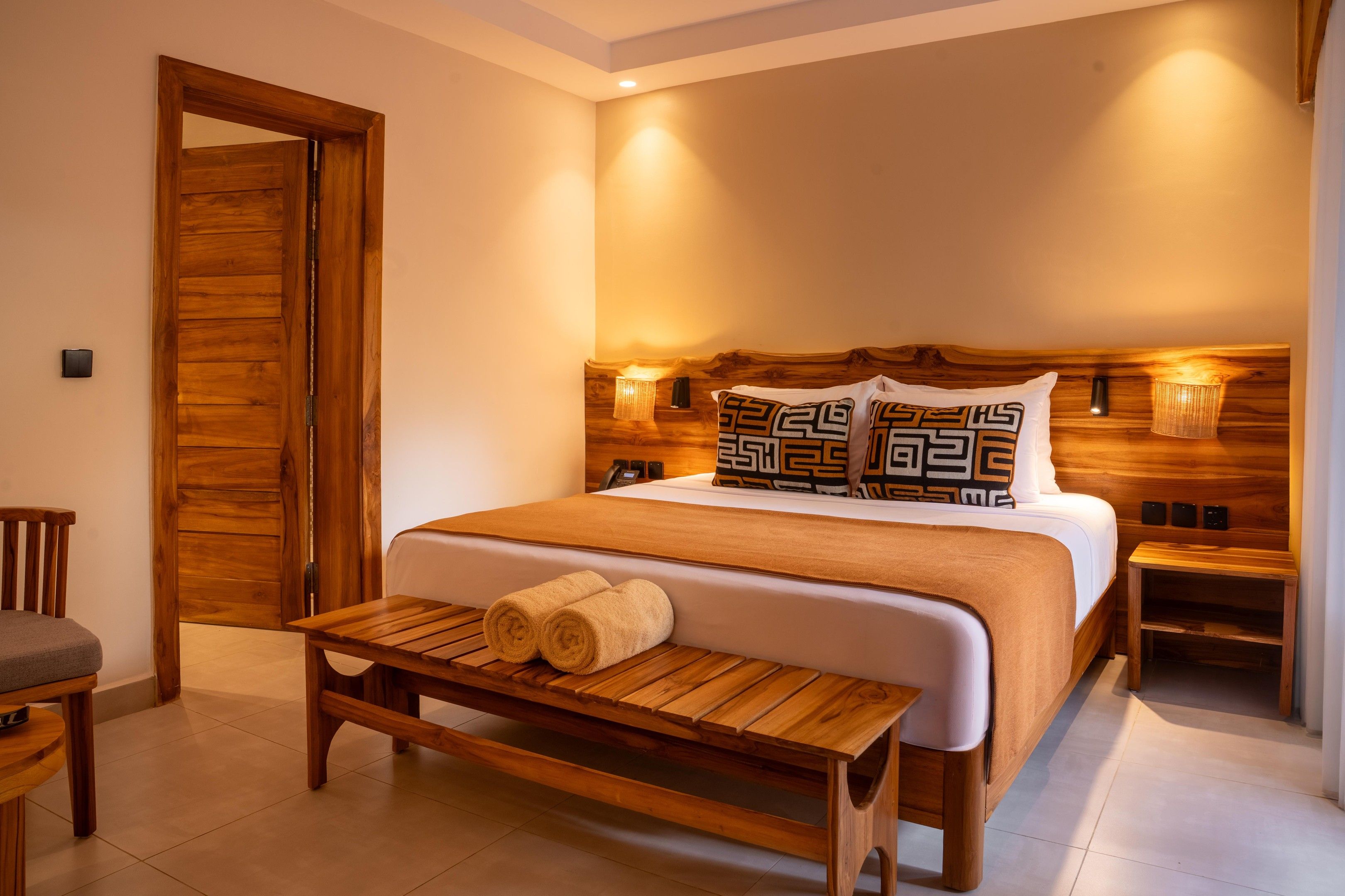 A hotel room with a wooden door, a bed with patterned pillows, a bedside table, a wooden bench with towels, and a small bedside table with a lamp.