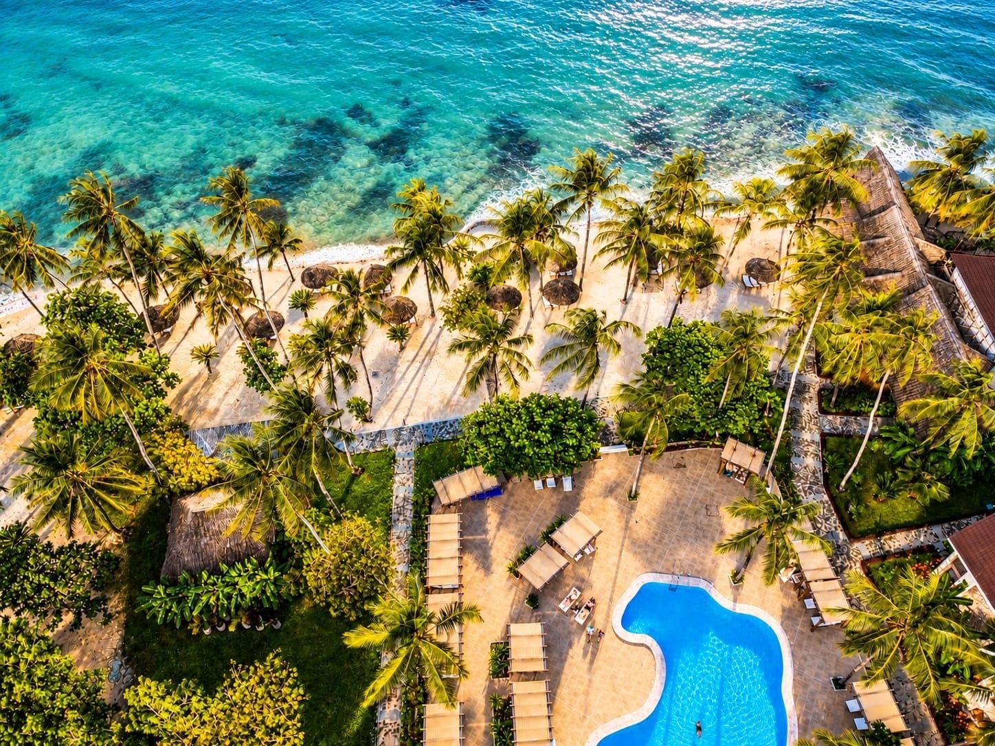 An aerial view of a resort with a swimming pool, palm trees, and a beach.