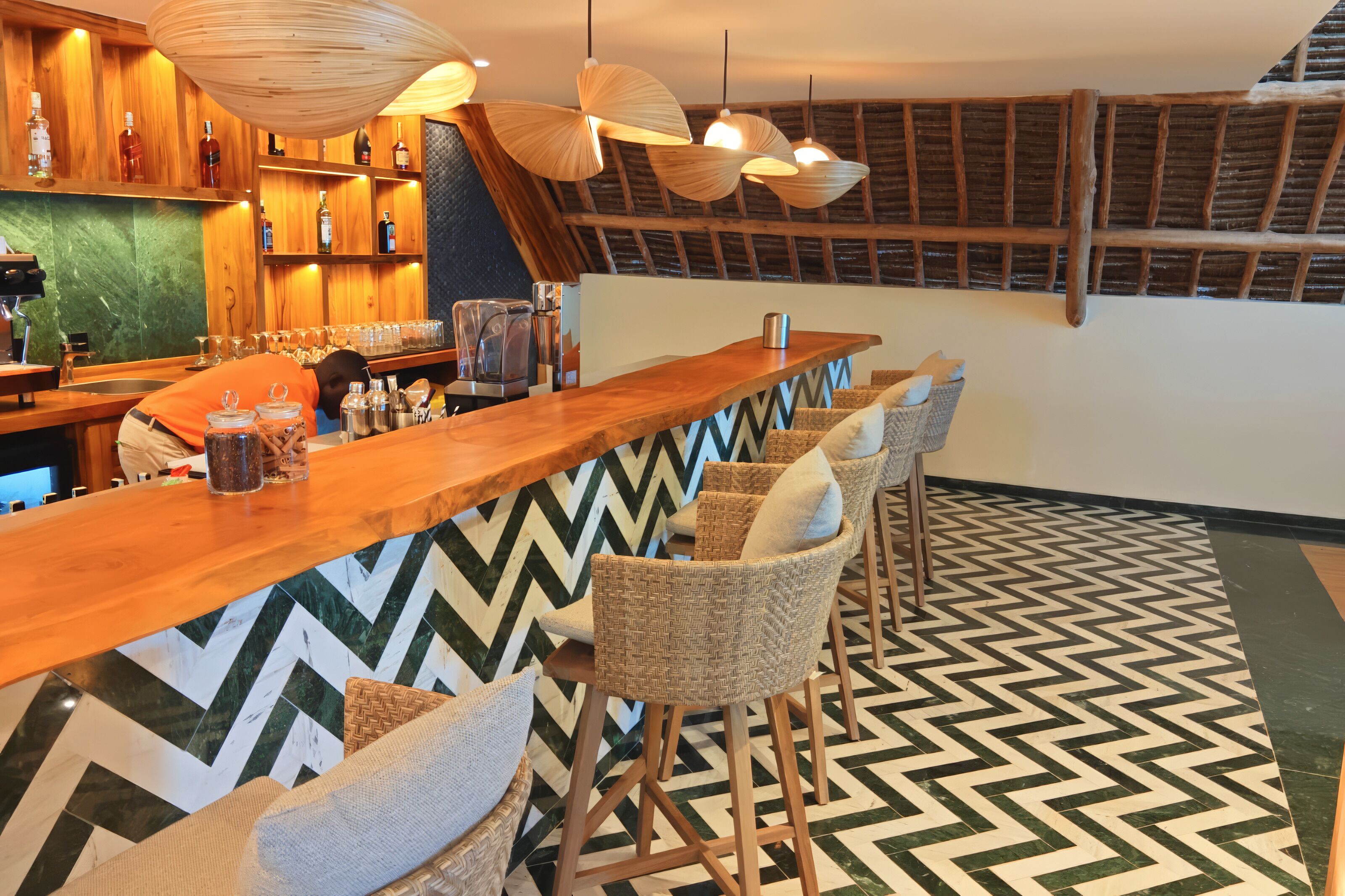 A bar area with wooden bar counter, wicker chairs, hanging light fixtures, and a chevron-patterned floor.