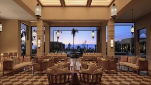 Un salón amplio con piso a cuadros, muebles de mimbre y grandes ventanas que ofrecen vistas a palmeras y una cadena montañosa distante.
