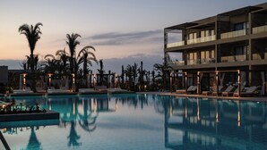 Zona de piscina con sillones reclinables, palmeras y un edificio moderno con balcones.