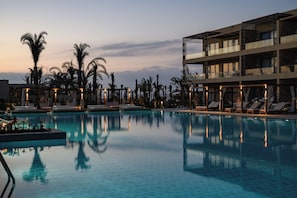 A pool area with lounge chairs, palm trees, and a modern building with balconies. - Sakhra Hotel & Spa, Almaza Bay (Marsa Matrouh)