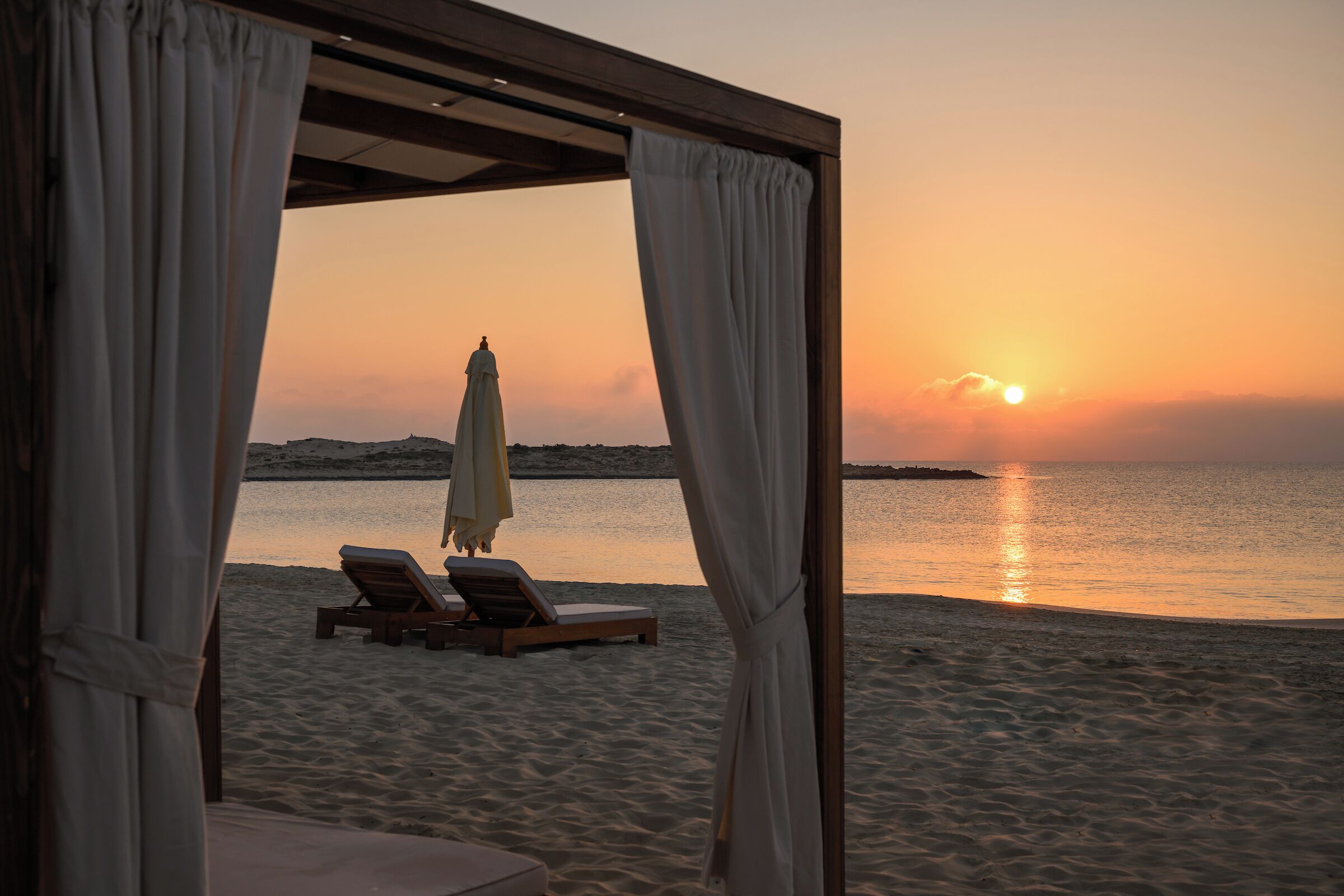 Een uitzicht op het strand met een zonsondergang, een beschaduwd zitgedeelte met een stoel en parasol, en een zandstrand.