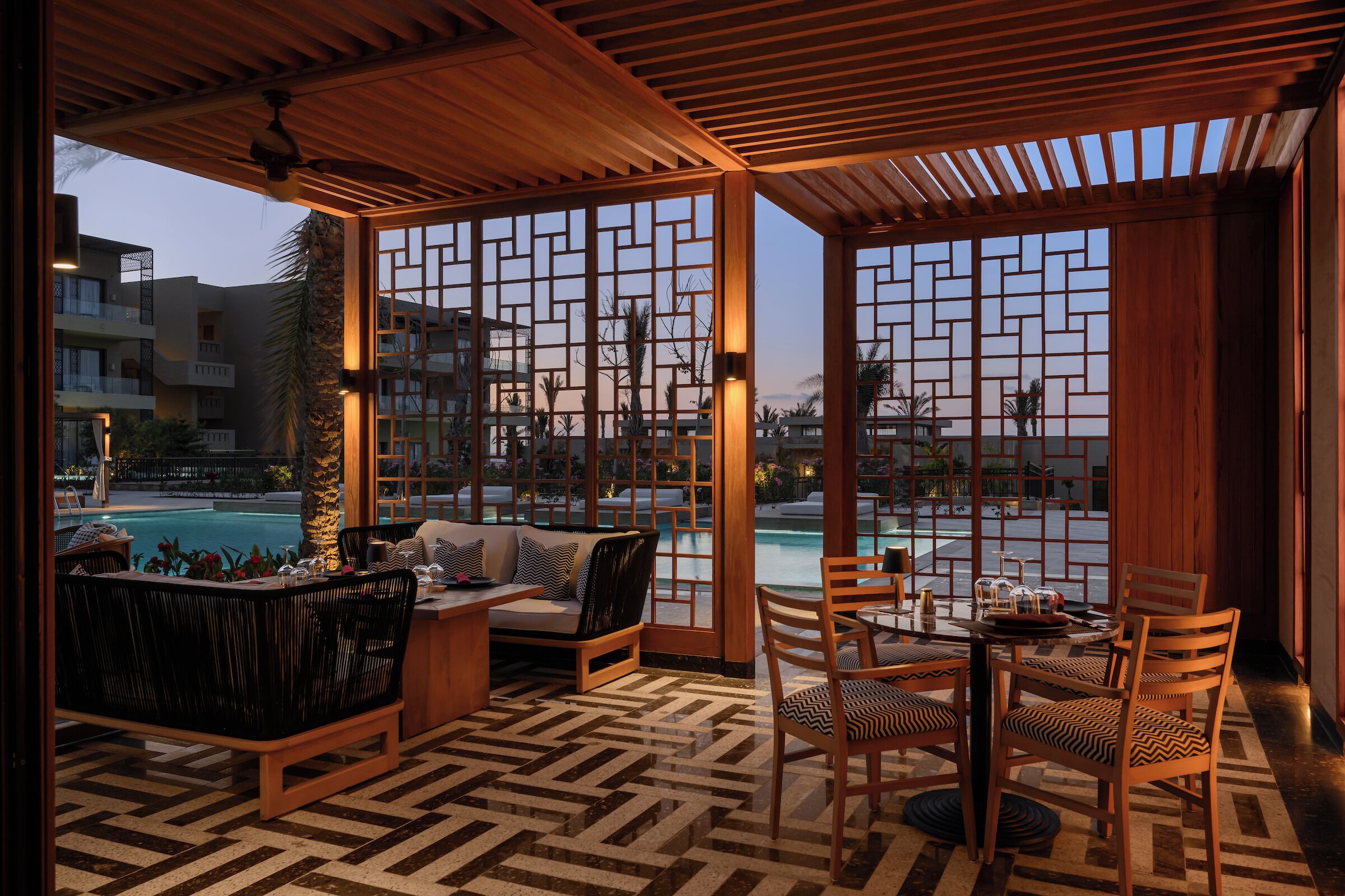 Un área de estar al aire libre cubierta con vista a la piscina, muebles de madera y piso con diseño.