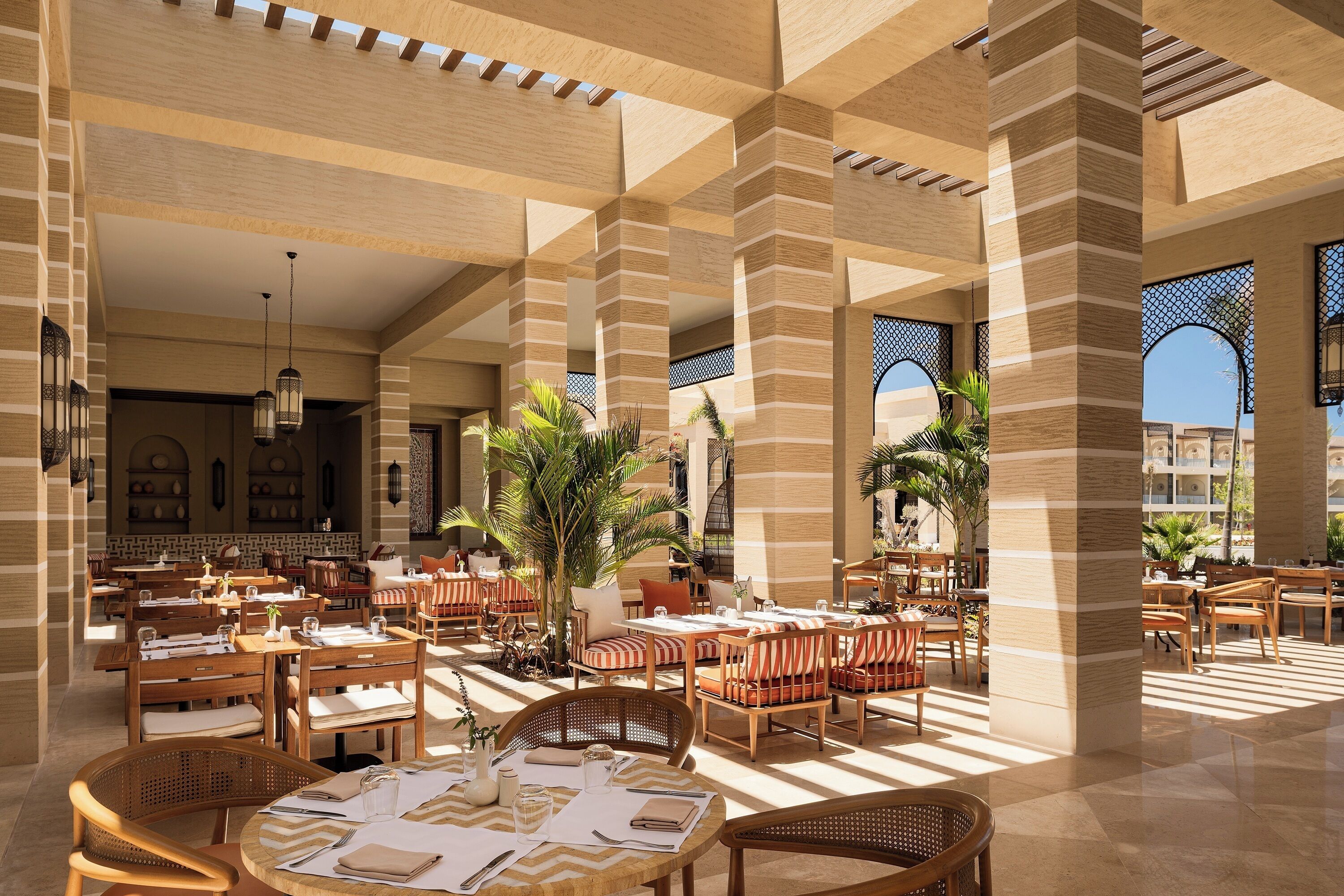 A spacious outdoor dining area with wooden tables and chairs, surrounded by columns and potted plants.