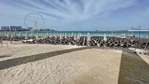A beach with rows of sun loungers and umbrellas, a Ferris wheel in the background, and a clear sky. - La Vie - Spacious One Bedroom (Jumeirah Beach Residence)