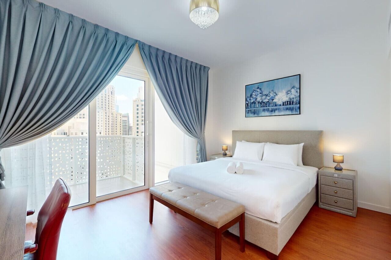 Quarto de hotel com uma cama grande, um banquinho, uma mesinha de cabeceira e vista para a cidade através de janelas do chão ao teto.