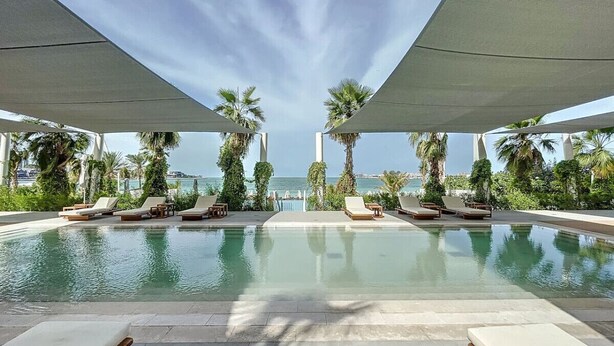 A pool area with lounge chairs and palm trees, overlooking a body of water. - La Vie - Spacious One Bedroom (Jumeirah Beach Residence)