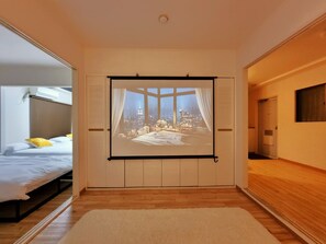 Un dormitorio con un ventanal grande, una cama y una pantalla de proyección que muestra un paisaje urbano.
