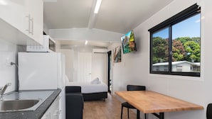 A compact kitchen with a refrigerator, microwave, and sink. There is a bed with a white headboard and a wooden table with chairs. A painting is visible on the wall, and a window offers a view of trees and a house.