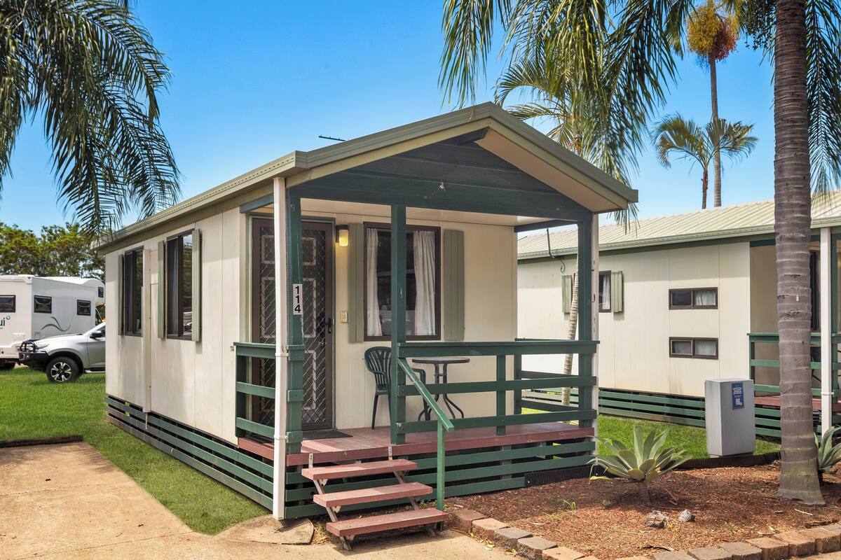 A small, single-story mobile home with a porch, a number '114' on the door, and a parked RV in the background.