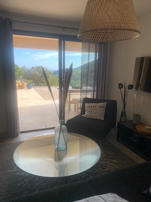 Ein modernes Wohnzimmer mit einem runden Glastisch, einem schwarzen Sofa, einer Korbtischlampe und Blick auf eine gebirgige Landschaft durch große Fenster.