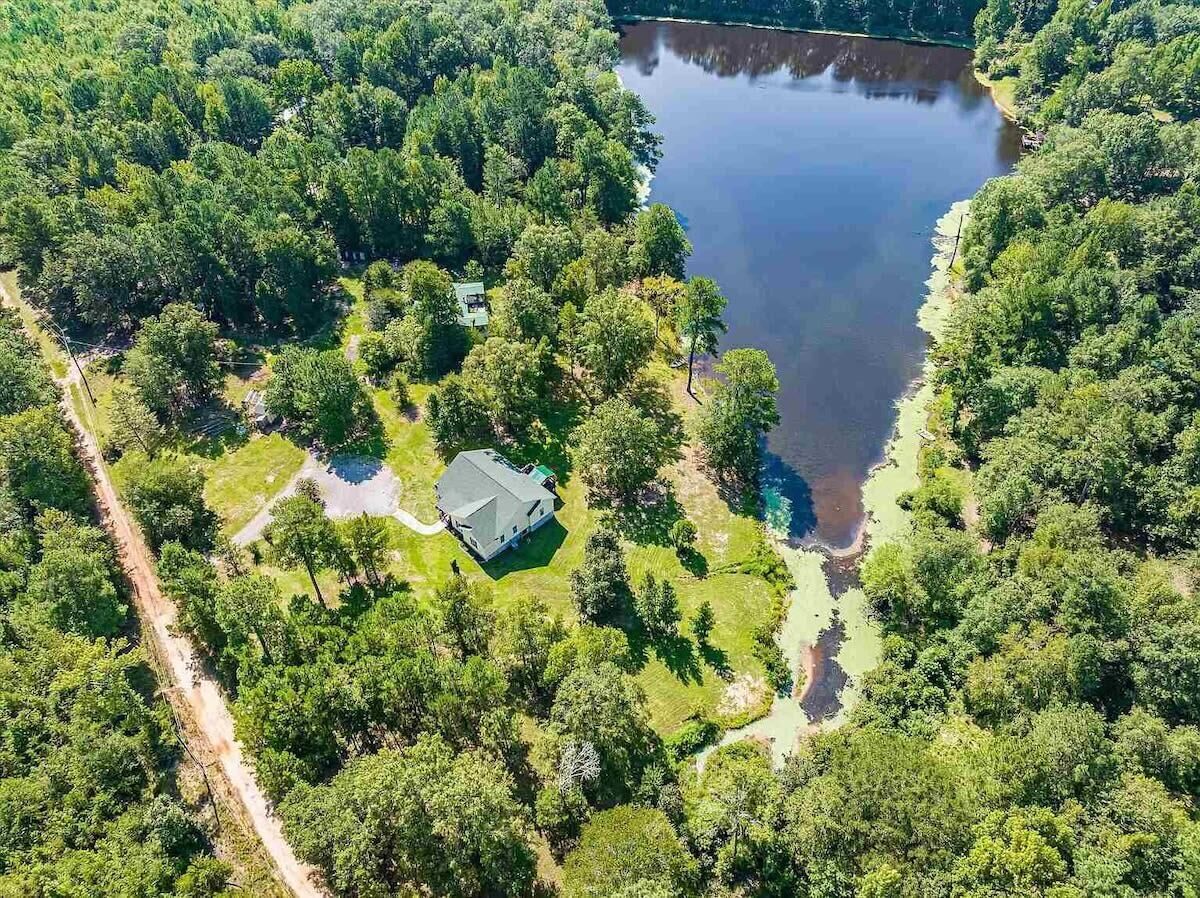 An aerial view of a property with a large lake, surrounded by dense forest. There is a house with a fenced yard and a small dock extending into the water.