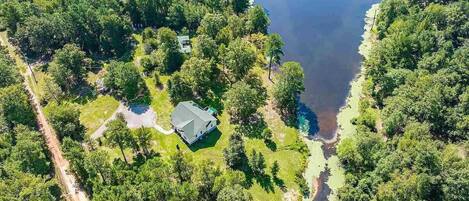 An aerial view of a property with a large lake, surrounded by dense forest. There is a house with a fenced yard and a small dock extending into the water.