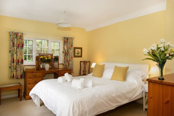 A bedroom with a large bed, a wooden dresser, a desk, and a chair. There is a window with curtains, a bedside table with a lamp, and a vase with flowers. - Woodcutter`s Cottage - Peaceful Country Retreat (Over Worton)