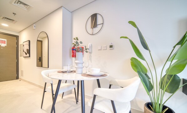 Quarto de hotel moderno com mesa de jantar redonda, cadeiras brancas, planta em vaso, relógio na parede e espelho.