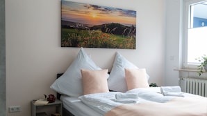 A neatly made bed with white and peach bedding, a bedside table with a book and a flower, and a painting of a sunset landscape on the wall.