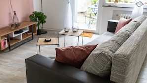 A modern living room with a black sofa, a wooden side table, and a glass coffee table. There is a potted plant and a small table with decorative items.