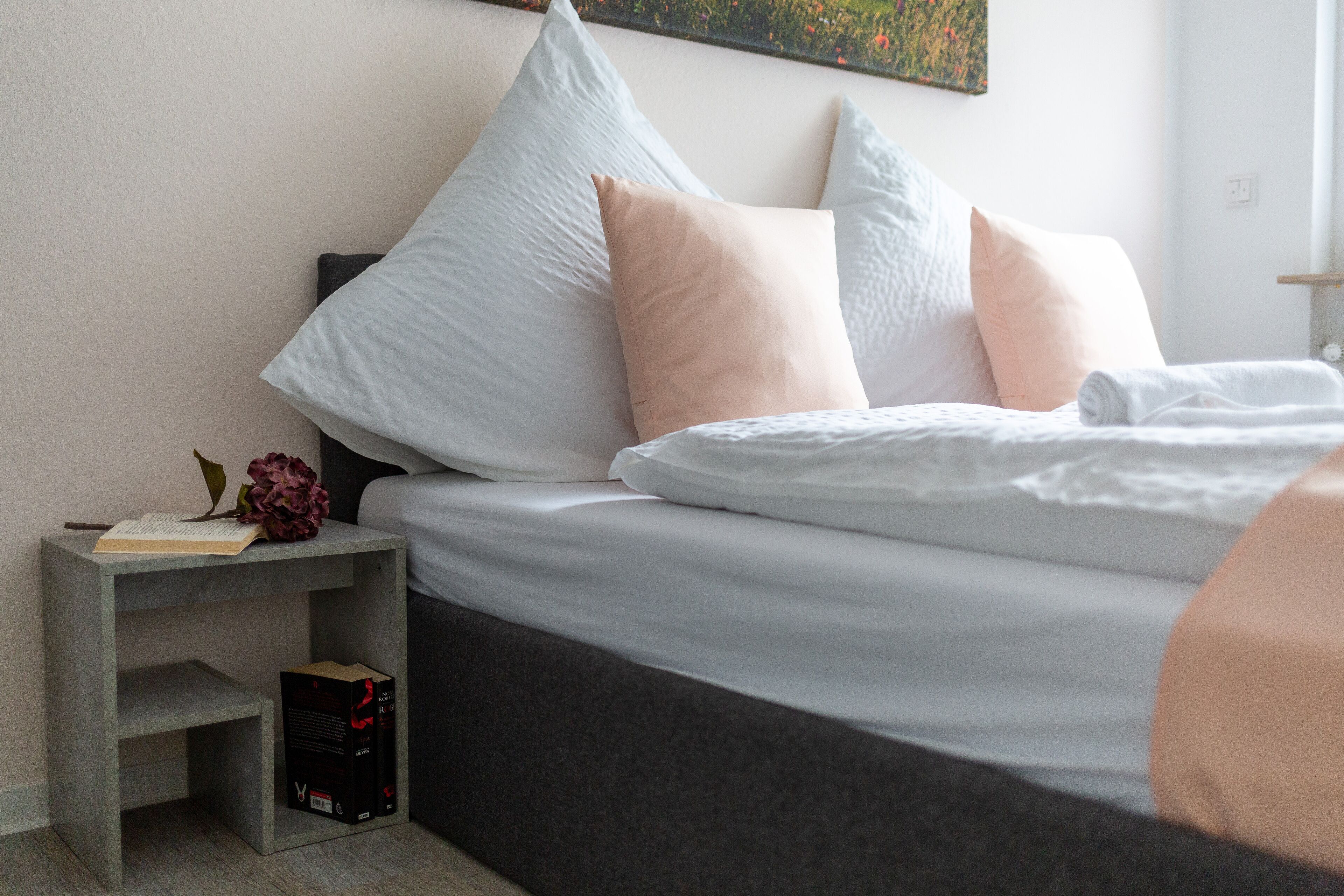 A neatly made bed with pillows, a bedside table with a book and a box, and a wall with a painting.